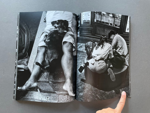 An open black-and-white photography book spread open on a gray surface, showing two street-style photographs printed on the pages. A person's hand is visible at the bottom right pointing toward the book.