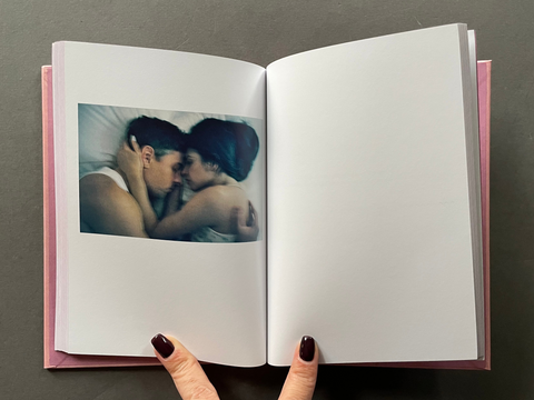 An open hardcover photo book with a pink cover, held open by two hands. The left page features a printed photograph of a couple embracing, while the right page is blank.