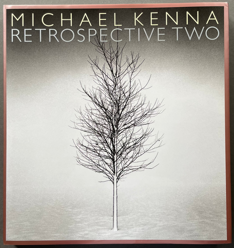 A square book or album cover titled 'Michael Kenna Retrospective Two' lies on a dark gray surface. The cover features a minimalist black-and-white tree illustration centered on a light gray background, framed by a muted reddish-brown border.