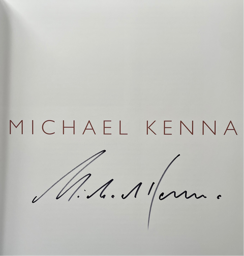 An open book or booklet showing a white page with the name MICHAEL KENNA printed in reddish-brown capital letters and a black ink signature below.
