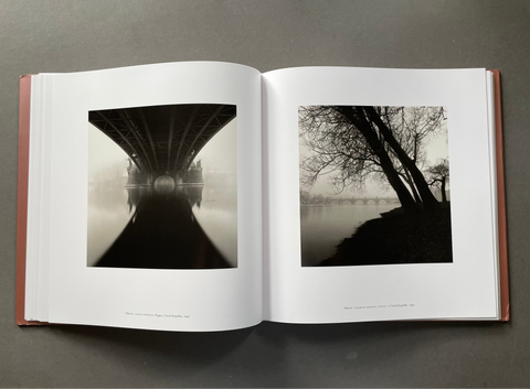 An open hardcover book lying flat on a dark gray surface, displaying two black and white landscape photographs printed on the pages: a bridge over misty water on the left, and bare trees beside a river on the right.