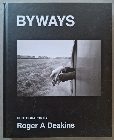 A hardcover book titled "BYWAYS" with a black cover, featuring a central black-and-white photograph of a hand resting on a train window beside a field. Text at the bottom reads "PHOTOGRAPHS BY Roger A Deakins.