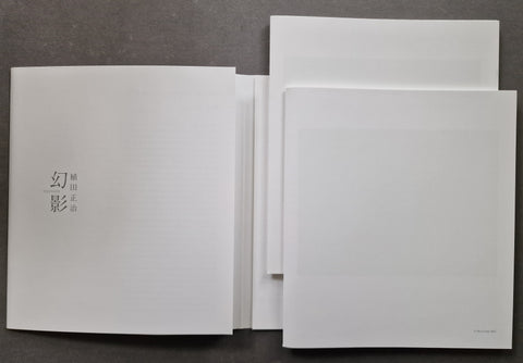 A stack of minimalist white art prints or booklets with Japanese text, arranged on a dark gray textured surface.