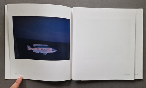 An open white art book on a dark gray surface, showing a printed photograph of a small white boat on dark blue water inside the left page