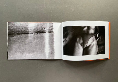 An open art book or photo journal laid flat on a gray surface, showing two black-and-white photographic prints side by side. The left page depicts a textured landscape or water scene, and the right page shows a close-up black-and-white photograph of a person's lower torso and hips.