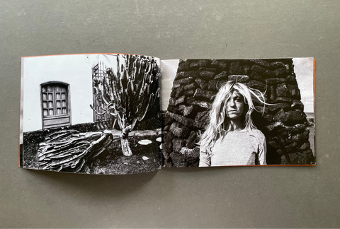 An open book or photo print spread laid flat on a gray surface, showing two black-and-white photographic scenes: a cactus and architectural detail on the left page, and a woman standing against a stone wall on the right page.