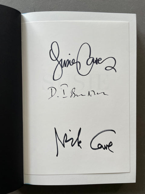 An open book or notebook showing a white page with three black handwritten signatures, photographed against a dark gray background.