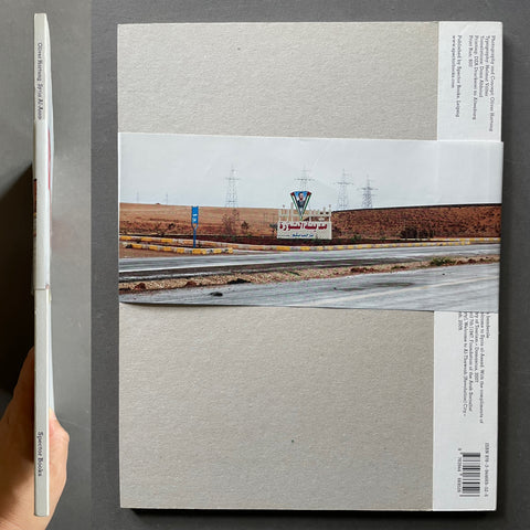 A square art print or poster with a large central landscape photograph of a highway and control tower, bordered by wide light gray textured paper sections and printed text blocks on the right and bottom edges. A hand is holding a matching slim copy of the print on the left against a dark gray background.