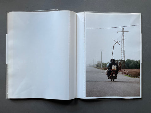 An open book or photo album laid flat on a dark gray surface, showing a large blank white left page and a printed photograph on the right page of a road scene with a person riding a motorcycle under overcast skies.