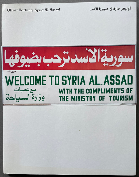 A book cover titled 'Welcome to Syria Al-Assad' with bilingual English and Arabic text, featuring a red banner and green lettering on a white background, photographed against a dark gray surface.