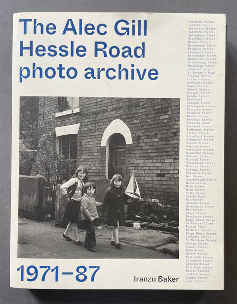 Book cover of 'The Alec Gill Hessel Road photo archive 1971-87' by Iaranzu Baker with a black and white photo of three children in front of a brick building.