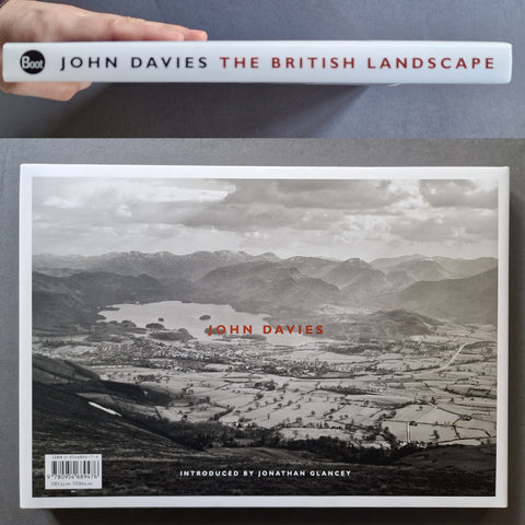 A hardcover book titled "John Davies The British Landscape" is shown from the front cover, held at the top by a hand. The cover features a black-and-white landscape photograph with red title text and a black publisher logo in the upper left.