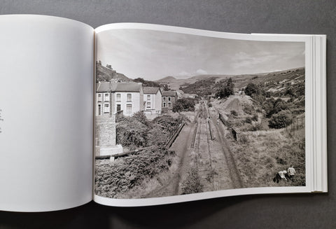 An open book showing a black-and-white landscape photograph print on the right page, depicting houses, hills, and railway tracks, photographed against a dark gray background.
