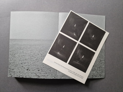 A black and white art poster lies open on a dark gray surface, with a smaller printed sheet placed on top showing four dark square scenes and small caption text.