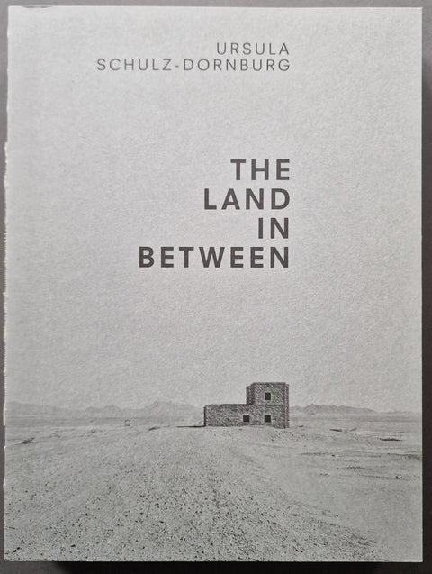 Book cover of 'The Land in Between' by Ursula Schultz-Dornburg, featuring a minimalist black and white photograph of a small stone structure in a vast desert landscape.