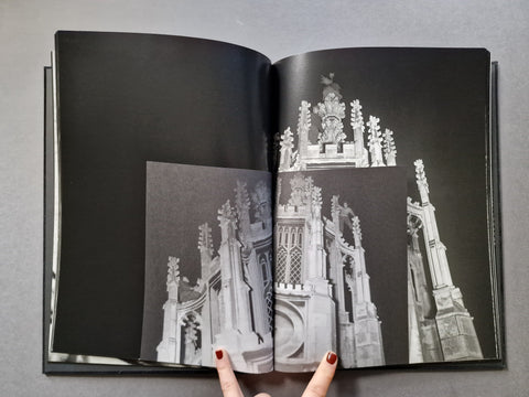 An open black-and-white architectural photo book with a hand holding an inset page showing detailed gothic building facades.