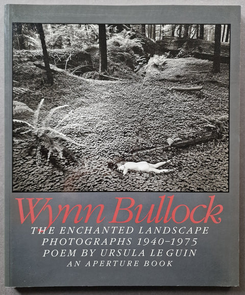 Book cover of 'Wynn Bullock: The Enchanted Landscape' featuring a black and white photograph of a forest path with a body on the ground, and red and white text on a gray background.