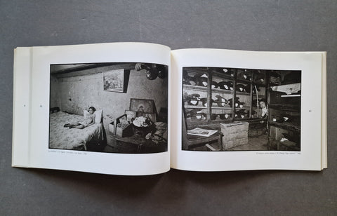 An open book laid flat on a dark gray surface, showing two black-and-white photographic prints on the pages. The left page contains a dim interior scene with a person lying on a bed, and the right page shows shelving with hats, with a small kid hiding in the background.