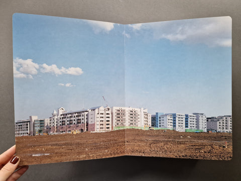 AN open book showing a construction site with a dirt foreground, a row of apartment buildings in the distance, and a blue sky with clouds.