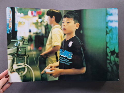 A printed photo book, showing a printed photograph of a young boy in a black graphic t-shirt playing with a game controller in a brightly lit arcade or gaming room.