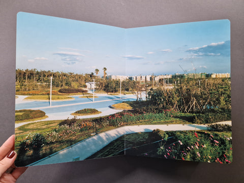 An open printed booklet showing a landscaped park scene with curved walkways, trees, flowers, and buildings in the distance under a blue sky.