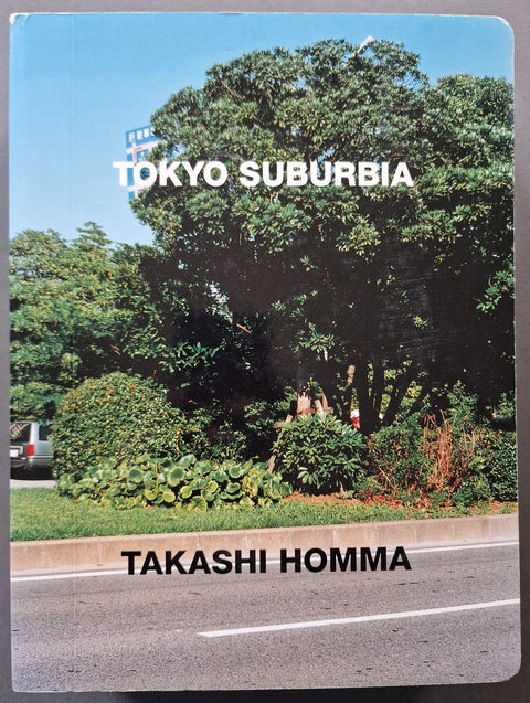 A book cover titled 'TOKYO SUBURBIA' by Takashi Homma, featuring a photograph of a lush green suburban street scene with trees, shrubs, and a road under a blue sky.