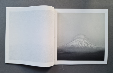 An open book showing a black and white photograph of a snow-capped mountain on the right page, with an empty blank page on the left, photographed on a dark gray surface.