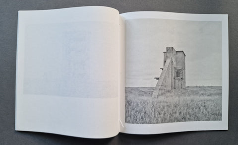 An open book showing a monochrome photograph of a tall industrial structure standing in a field, photographed from above on a dark gray surface.