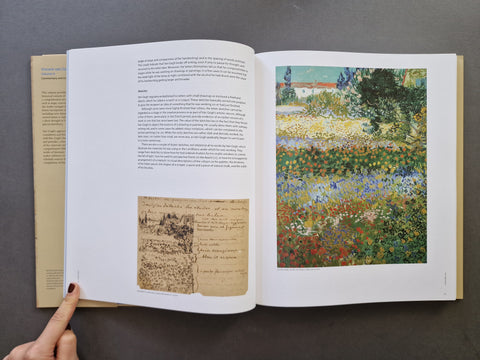 An open book laid flat on a dark gray surface, showing printed text on the left page and a colorful impressionist-style landscape painting on the right page. A person's finger is visible holding the left edge of the book.