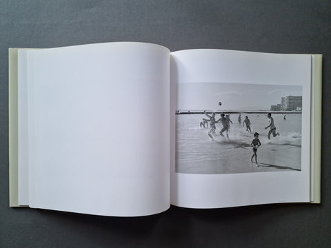 An open book, showing a black-and-white photograph of people playing in shallow water on the right page and blank white pages on the left.