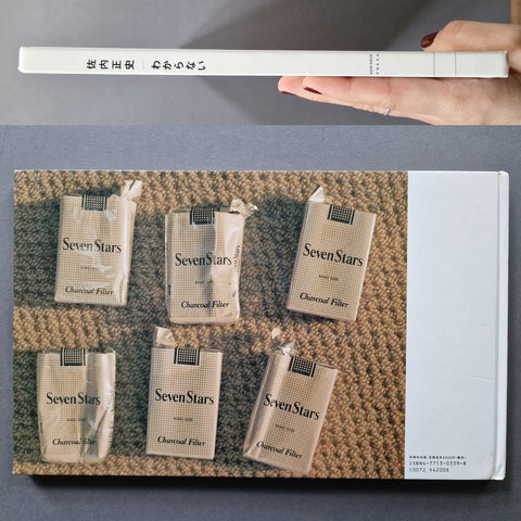 A hand holds a slim white book or booklet above a larger printed cover featuring five illustrated packs of SevenStars Charcoal Filters arranged on a textured brown surface. The smaller item shows the title 'SevenStars' and the larger cover includes the same branding and a white side panel.