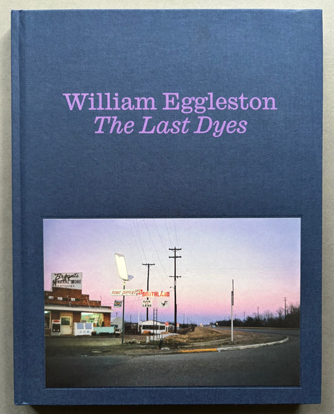 Book titled 'The Last Dyes' by William Eggleston with a blue cover featuring the image of a building on the corner of an American road.