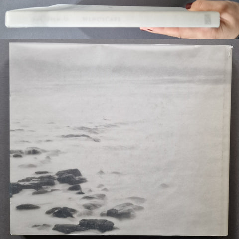 A large black and white photographic print or book cover featuring a seascape with rocks and misty water, held horizontally by a hand at the top edge against a dark gray background.