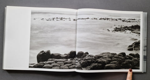 An open hardcover book showing a black and white photographic print of a rocky shoreline, photographed from above on a dark gray surface with a finger visible at the bottom right.