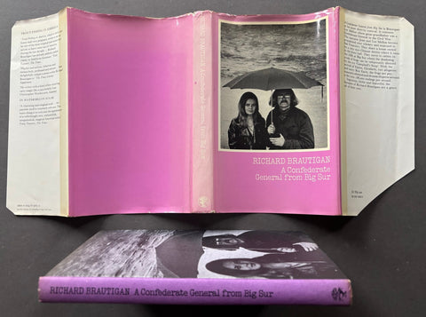An open vintage hardcover book titled 'Richard Brautigan: A Confederate General from Big Sur' displayed above its closed cover, both resting on a dark surface. The book's cover and interior pages feature a prominent pink background with a black-and-white photograph of two people under an umbrella.