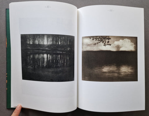 An open book showing two printed landscape photographs on the pages: a black-and-white forest reflection on the left page and a sepia-toned cloudy sky over water on the right page. A finger is visible holding the left page at the bottom.