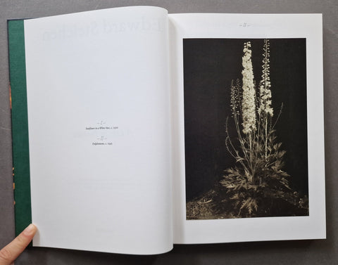 An open book showing a printed page with a black-and-white botanical photograph of tall flowering stems and a dark green spine, held by a hand against a dark gray background.