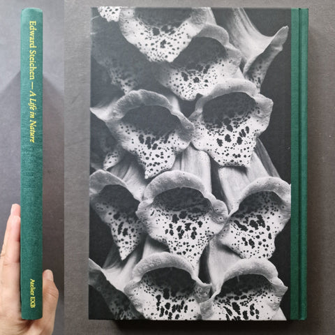 A hardcover book titled 'Edward Seiden - A Life in Nature' by Adler & Co, displayed upright next to a hand holding its green spine to show scale. The cover features a dramatic black and white macro photograph of foxglove flowers, with a dark green spine and matching green binding.