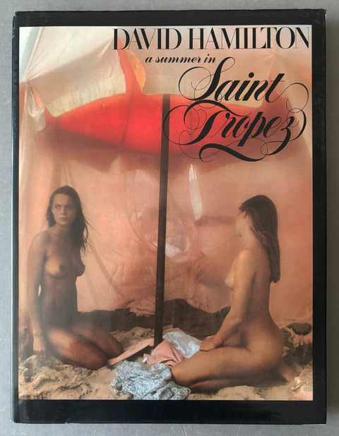 A vintage hardcover book titled 'A summer in Saint Tropez' by David Hamilton, featuring a cover photograph of two nude women seated under a red beach umbrella on a sandy beach.
