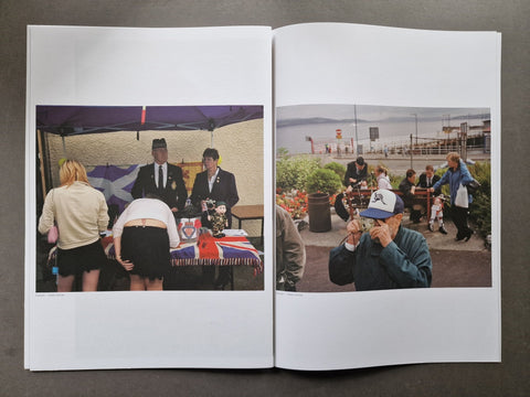 An open publication laid flat on a dark surface, showing two printed photographs side by side. The left page contains a photo of people gathered under a canopy around a table with a Union Jack cloth.