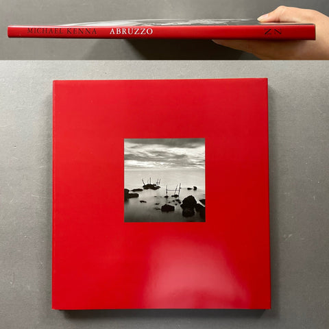 A hardcover art book titled 'Michael Kenna Abruzzo' is shown, with the top half displaying the red spine and the bottom half showing the red cover with a central black-and-white photograph of rocks in shallow water. A hand is visible holding the spine at the top edge.