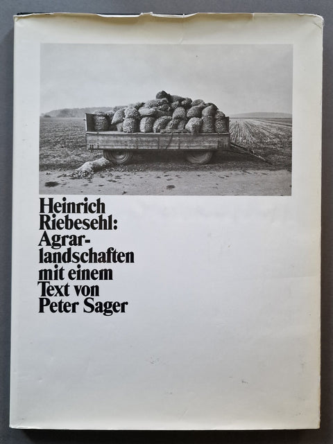 A book cover for 'Heinrich Riebeschl: Agrar-landschaften mit einem Text von Peter Sager' featuring a black and white photograph of a cart loaded with sacks on a dirt road, set against an off-white background.