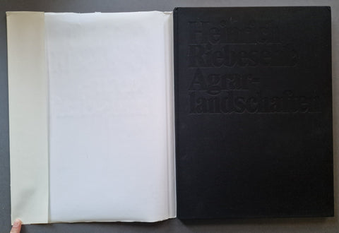 An open book showing a large black embossed cover with the title 'Heinrich Riebesehl Agrarlandschaften' and a white inner page or folder beside it, photographed against a dark gray surface.
