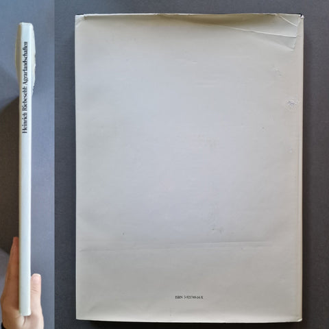 A hand holds a slim white book or art print next to a larger off-white square sheet or box on a dark gray background. The spine shows the text 'Heinrich Riebesehl Agrarlandschaften' and the bottom of the square piece shows 'RIN.