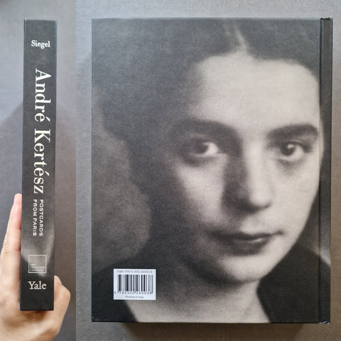 A hand holds a slim book spine next to the full book cover of 'Postcards from Paris' by André Kertüsz, published by Yale. The cover features a black and white portrait of a woman, with the spine showing the title and Yale publisher branding.