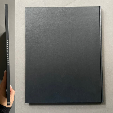 A hardcover art book or photo album with a dark charcoal gray textured cover, shown from the front and spine. A hand is holding the spine on the left to show the thickness.