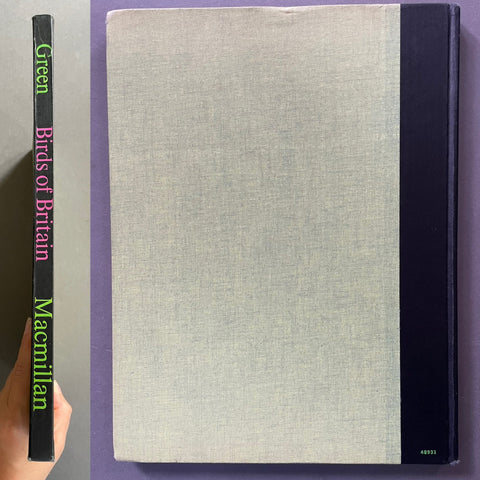 A hardcover book titled 'Green Birds of Britain' by Macmillan, held by a hand against a purple background. The book features a light gray linen-textured front cover, a black spine with colorful title text, and a black binding strip on the right side.