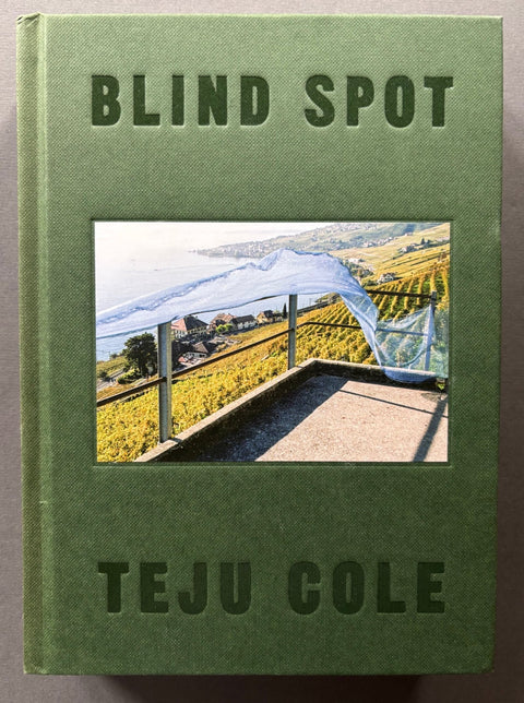 A hardcover book titled 'BLIND SPOT' by Teju Cole, featuring an olive green textured cover with a central photograph of a vineyard landscape under a white canopy, set against a dark gray background.