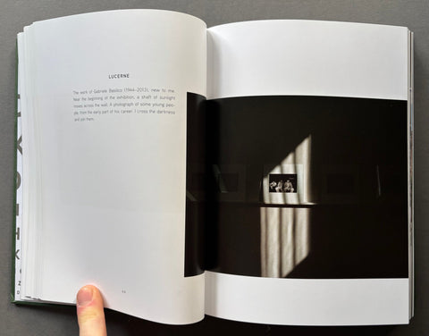 An open book or art print showing a two-page spread with text on the left and a black-and-white photograph on the right. A human finger is visible holding the left page at the bottom.
