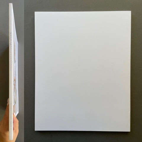 A white book shown flat against a dark gray background, with a hand holding the left edge to show the spine and thickness of the print.